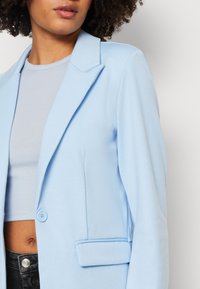 Blazer bleu clair en tissu doux, doté d'un col cranté, d'un bouton unique et d'une poche latérale. Associé à un haut gris côtelé.