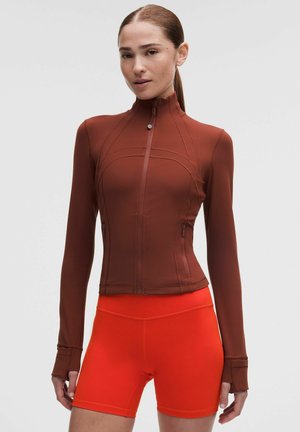 Kvinde iført en tætsiddende brun lynlåstrøje med tommelfingerhuller og klare orange højttaljede cykelshorts mod en ensfarvet grå baggrund.