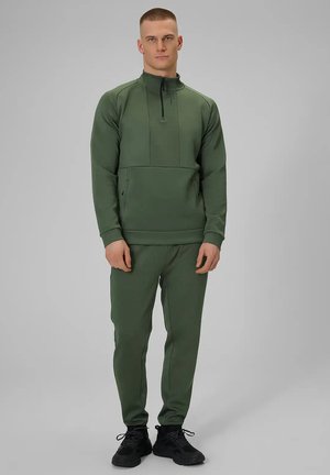 Man staand, draagt een olijfgroene zip-up sweat en bijpassende joggerbroek met zwarte sneakers op een grijze achtergrond.