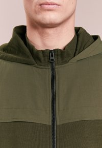 Olivengrøn hættejakke med lynlås foran, der har kontrasterende teksturmønstre, en tætsiddende hals og synlige syningsdetaljer.