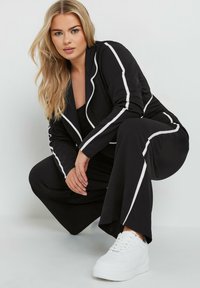 Czarna marynarka i spodnie z białymi paskami po bokach, w połączeniu z białymi sneakersami. Gładki materiał i dopasowany krój. Model klęczy.