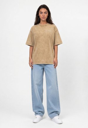 Vrouw staand, dragend een losse beige korte mouwen blouse, lichtblauwe wijde jeans en witte sneakers tegen een effen witte achtergrond.