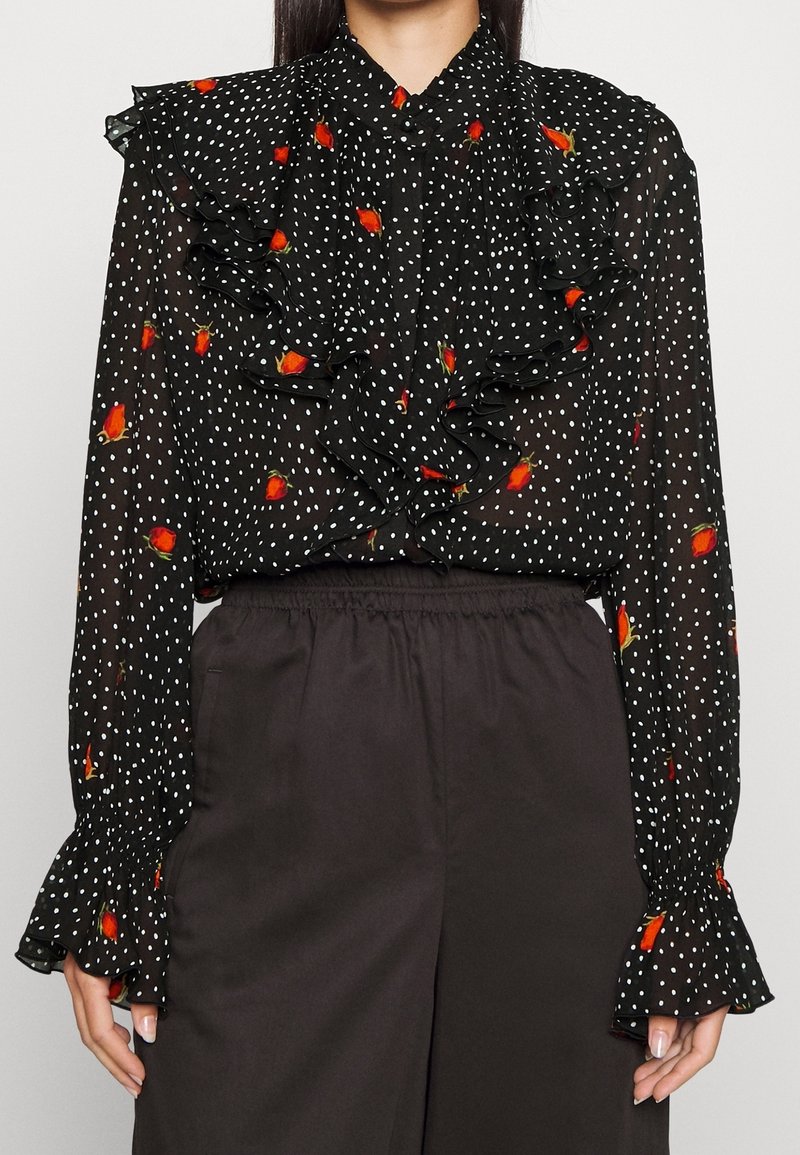Blouse noire transparente à pois blancs, boutons de rose rouges éparpillés, devant et poignets froncés, associée à un pantalon noir taille haute.