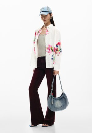 Vrouw draagt een witte bloemenblouse, grijze tanktop, bordeauxrode broek, spijkerpet, blauwe sandalen en houdt een blauwe Desigual tas vast.