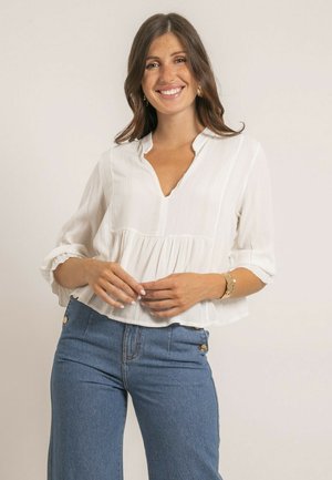 Blouse blanche texturée avec un col en V et des manches 3/4, présentant un design froncé à la taille ; portée avec un jean bleu taille haute.