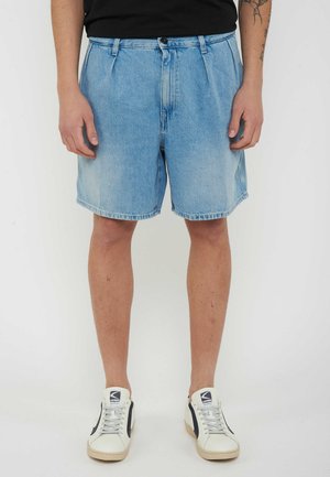 Uomo che indossa pantaloncini di jeans azzurri, camicia nera, sneakers bianche con dettagli neri, in piedi su sfondo bianco.