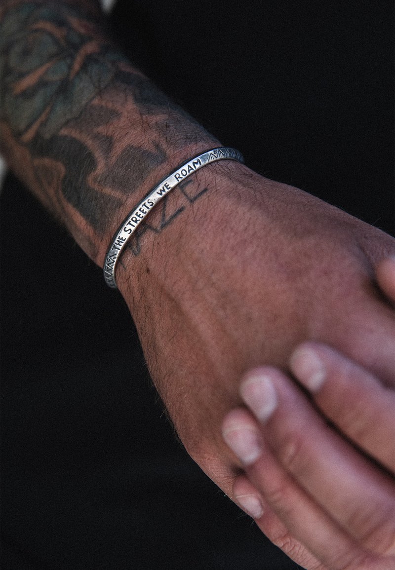 Metallarmband med ingraverad text "DE GATOR VI VANDRAR," med en slät, polerad yta och geometriska detaljer längs kanten.