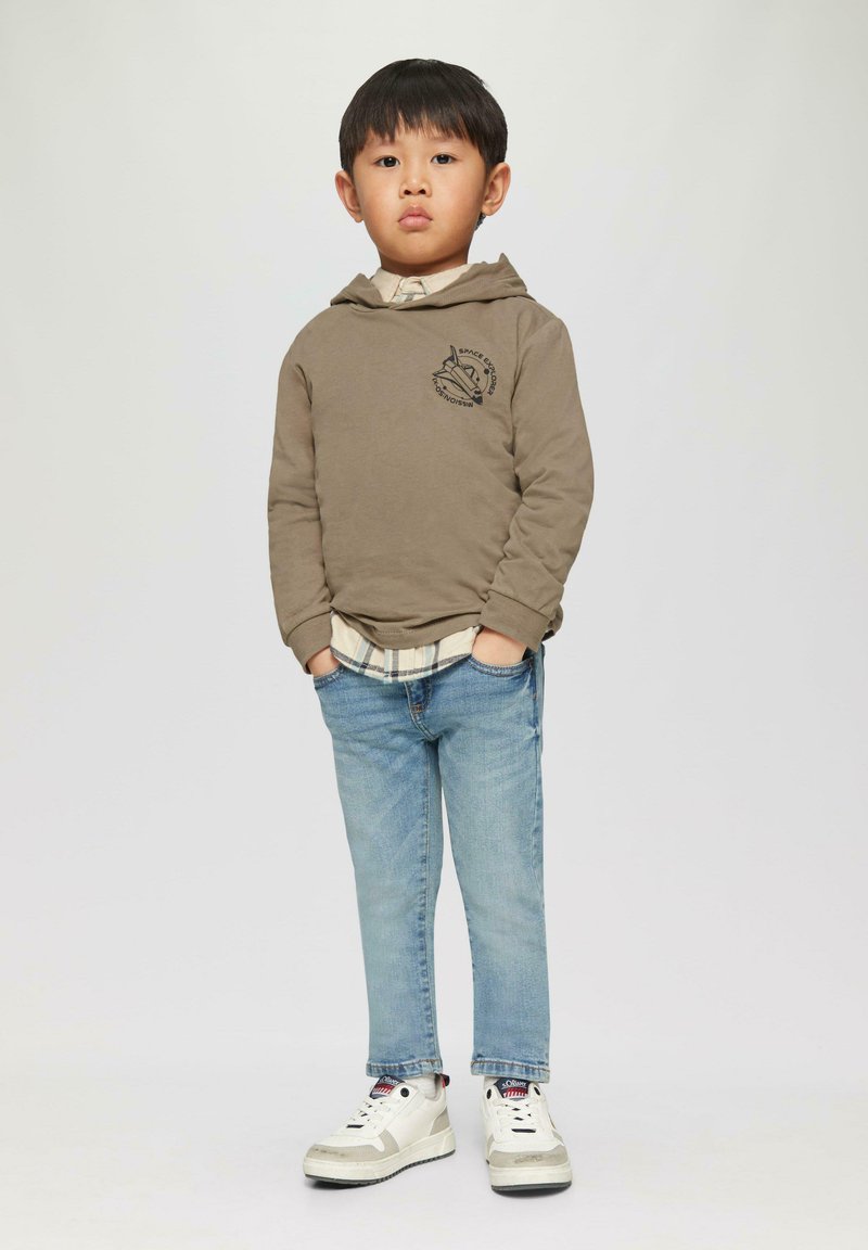 Khaki hoodie met logo, gelaagd over een geruite blouse, gecombineerd met lichtblauwe spijkerbroek en witte sneakers met grijze accenten. Eenvoudige, casual outfit.