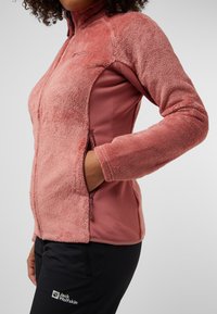 Fleecejacke in zartem Pink mit strukturierter Außenseite, hohem Kragen und seitlichen Taschen. Kombiniert mit schwarzen Hosen, die ein Logo haben.