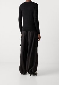 Personne portant un haut noir à manches longues et un pantalon cargo noir ample avec cordons aux chevilles, debout pieds nus sur un fond blanc.