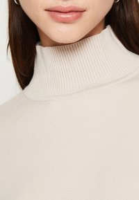 Bežani rebrasti turtleneck ovratnik s glatkom teksturom. Tkanina je lagana i prilagođena, naglašavajući područje vrata i brade.