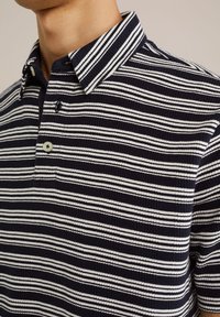 Jūras spēku un baltas svītras īsu piedurknu polo krekls ar pogājamu apkakli un faktūru. Iezīmēts ar vienu pogu pie kakla.