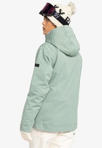 Lys grøn skijakke med hætte, elastiske manchetter og sidelommer. Sammen med en mønstret hvid strikbeanie og skibriller.