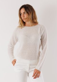 Donna con capelli castano chiaro lunghi fino alle spalle, che indossa un maglione bianco lavorato a maglia e pantaloni bianchi, in posa con le mani in tasca, che guarda di lato.