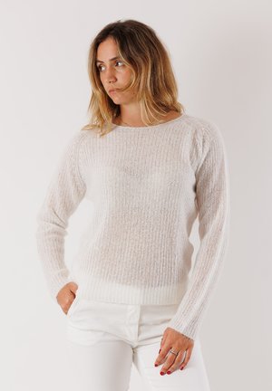 Donna con capelli castano chiaro lunghi fino alle spalle, che indossa un maglione bianco lavorato a maglia e pantaloni bianchi, in posa con le mani in tasca, che guarda di lato.