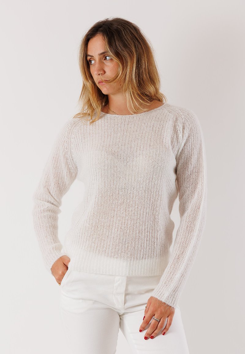 Donna con capelli castano chiaro lunghi fino alle spalle, che indossa un maglione bianco lavorato a maglia e pantaloni bianchi, in posa con le mani in tasca, che guarda di lato.