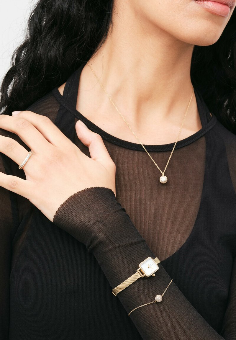 Gold watch with mesh band, layered gold necklace with a spherical pendant, and a thin gold bracelet. Black sheer long-sleeve top.