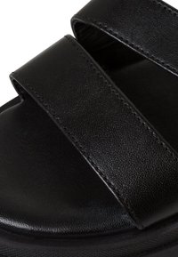 Black leather sandal with two wide straps and a textured sole edge, showing close-up details of the material and stitching.