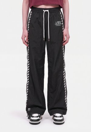 Una modelo lleva unos pantalones deportivos negros de pernera ancha, con cordones blancos, una franja lateral a rayas y un logotipo, combinados con zapatillas deportivas blancas y negras.