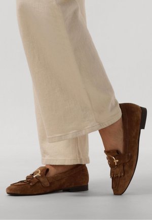 Mocassins en daim marron avec pompons et détail de boucle dorée portés avec un pantalon beige à jambes larges sur un fond clair.