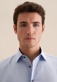 Camicia azzurra chiara con colletto classico, bottoni scuri e una consistenza liscia. Nessun motivo o logo visibile.