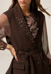 Donkerbruine vest met een gerimpelde bloemenblouse eronder. Voorzien van decoratieve studs op de kraag, een strikceintuur en twee voorzakken.