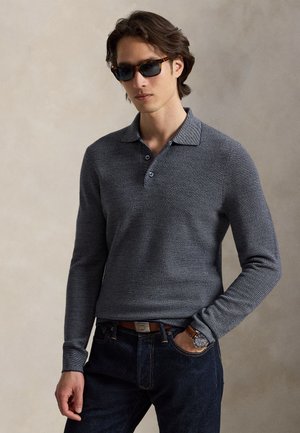 Mand iført mørke solbriller, en grå tekstureret langærmet polo, mørke jeans og et armbåndsur, med den ene hånd i lommen.