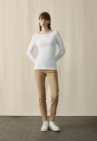 Vit långärmad topp med texturerad yta, tillsammans med ljusbruna byxor och vita sneakers. Modellen poserar med händerna bakom ryggen.