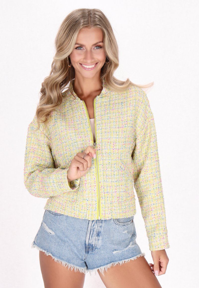 Geel tweed jasje met pastelkleuren, ronde kraag en ritssluiting aan de voorkant, gecombineerd met versleten denimshorts.