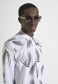 Camisa blanca con un diseño de hojas negras, que cuenta con un cuello tipo botonadura. El modelo lleva unas gafas de sol transparentes y angulares.
