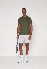 Oliven grøn T-shirt, grå shorts, holder en tennisracket og bold. Hvide sportssko med marineblå accenter og hvide sokker. Enkelt design.