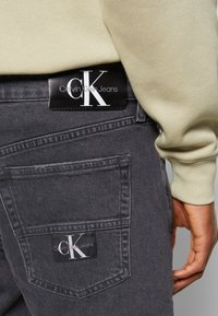Jeans gris oscuro con un acabado texturizado. Las características incluyen un bolsillo trasero con una etiqueta negra rectangular que muestra el logotipo "Calvin Klein Jeans".