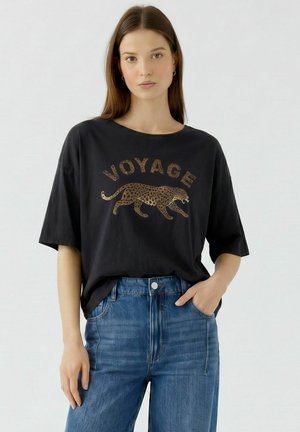 Jeune femme portant un t-shirt noir avec un léopard marchant et le mot "VOYAGE", associé à un jean bleu, debout devant un fond uni.