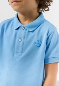 Lichtblauw poloshirt van gestructureerde stof. Heeft een twee-knopenbies en een rond logo op de linkerborst. Korte mouwen.