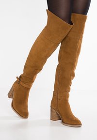 Hnědé semišové kozačky nad kolena se širokým ohybem, zipovým uzávěrem a detailem přezky. Blokový podpatek s jemnými vertikálními drážkami.