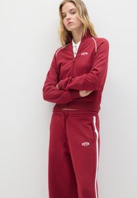 Giacca rossa con zip, dettagli bianchi e logo "BOSTON", abbinata a pantaloni felpati rossi con strisce bianche laterali. Tessuto morbido.