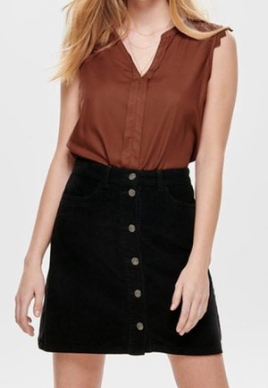 Mujer con blusa marrón sin mangas metida dentro de una falda negra evasé con botones al frente, de pie frente a un fondo liso.