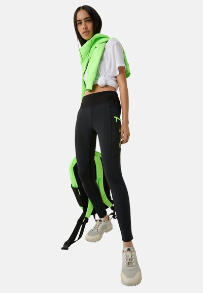 Schwarze Leggings mit hohem Bund, ausgestattet mit einer Reißverschlusstasche und neon-grünen Akzenten. Getragen mit einem weißen, geknoteten Shirt und einem über die Schultern drapierten grünen Sweatshirt.