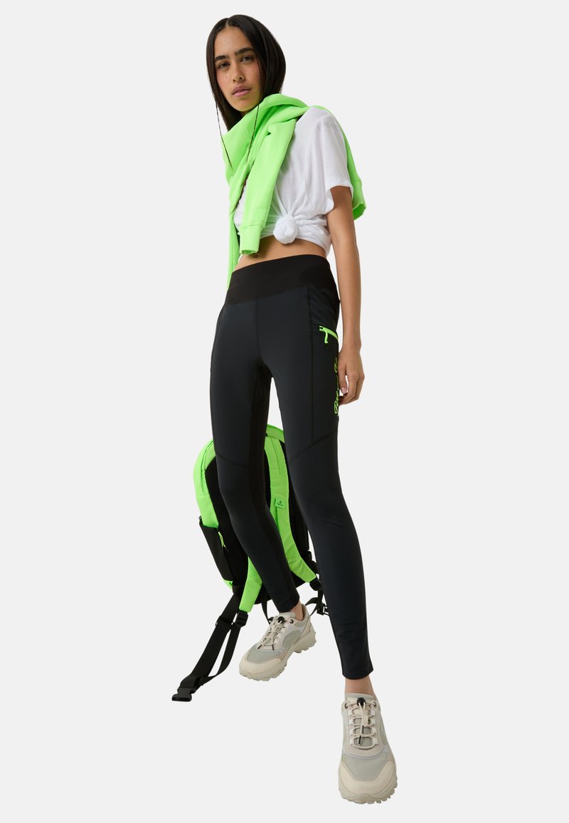 Schwarze Leggings mit hohem Bund, ausgestattet mit einer Reißverschlusstasche und neon-grünen Akzenten. Getragen mit einem weißen, geknoteten Shirt und einem über die Schultern drapierten grünen Sweatshirt.