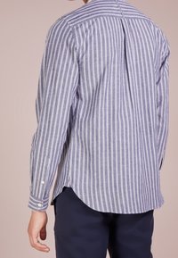 Camicia a righe blu e bianca in tessuto; presenta maniche lunghe, una piega sul retro e un orlo arrotondato con bottoni bianchi a contrasto.
