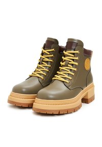 Botas tobilleras de color verde oliva con cordones amarillos, detalles marrones, suela gruesa de color beige y un logo circular en el lateral.