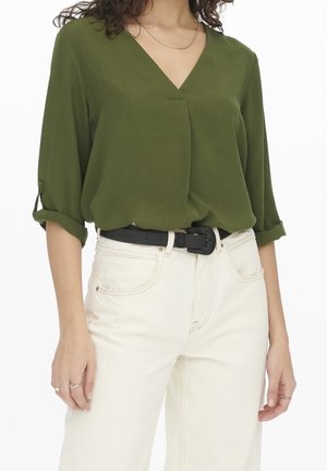 Woman wearing olive green V-neck blouse with rolled-up sleeves, white high-waist jeans, black belt, and gold chain necklace.