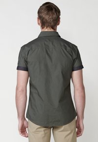 Camisa de manga corta en tela verde oscuro con un canesú trasero y puños enrollados. Detalles de costura y un dobladillo curvado.