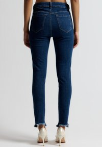 Tmavomodré úzke rifle s rozstrapkaným lemom, majú zadné vrecká a vysoký pás, vyrobené z denimového materiálu.