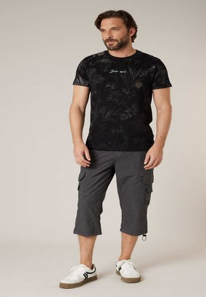 Homme portant un t-shirt noir à imprimé floral, un short cargo gris et des baskets blanches, debout devant un fond beige uni.