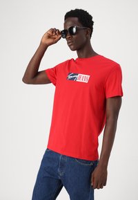 Rød bomuld t-shirt med et "Tommy Jeans" logo i navy og hvid, parret med mørkeblå jeans og sorte solbriller.