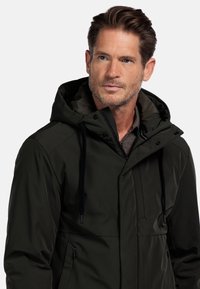 Donkergroene waterdichte jas met capuchon, voorkant met rits, verstelbare trekkoorden en zijzakken. Zachte textuur met een gestructureerd ontwerp.