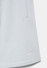 Shorts en tissu gris clair avec poche latérale et petit logo de marque brodé blanc près de l'ourlet sur un fond uni.
