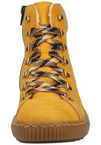 Botte montante en daim jaune avec des lacets à carreaux noirs et jaunes, quincaillerie dorée et semelle en caoutchouc texturée. Fermeture éclair sur le côté.