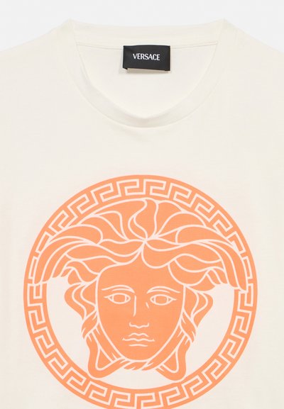 T-shirt blanc avec un visuel tête de Méduse orange entouré d'un motif clé grecque et une étiquette Versace noire cousue à l'intérieur de l'encolure.